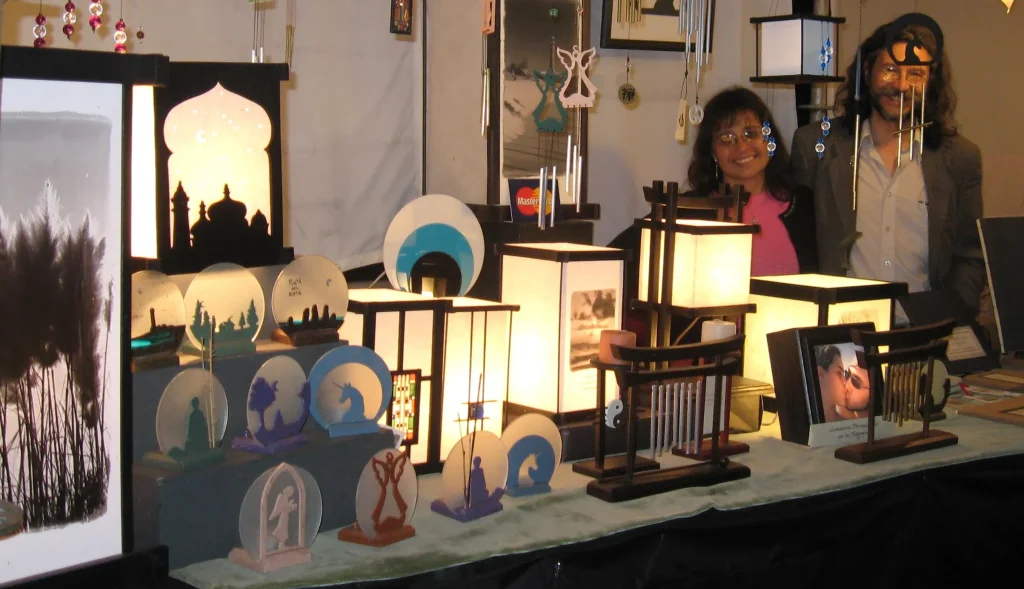 Display booth with illuminated lanterns and decorative sculptures, people smiling in the background. - Informative: a craft fair stall showcasing lit paper lanterns and silhouettes.