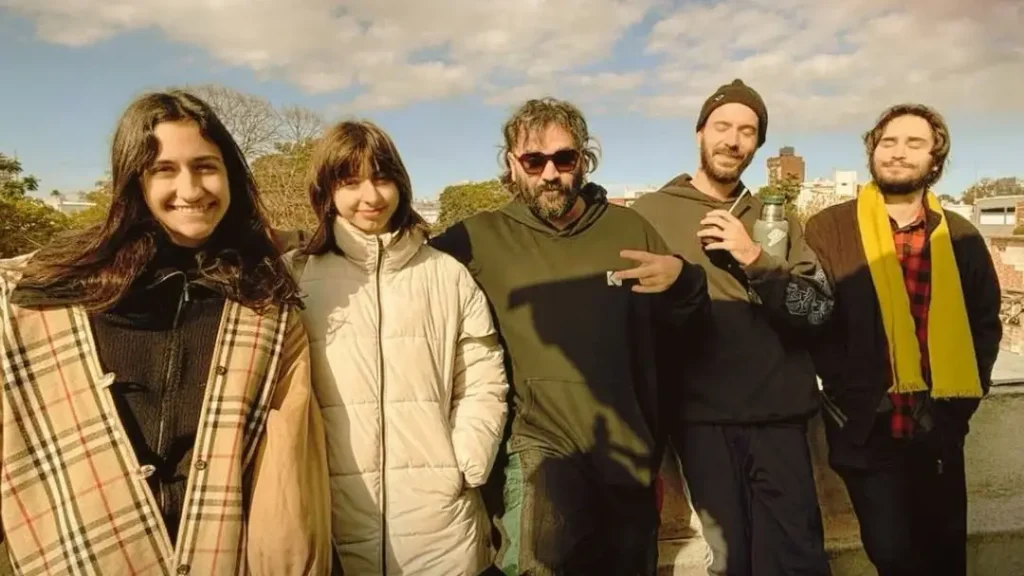 Five friends posing on a sunny rooftop, smiling at the camera in warm jackets and coats. Five people are standing together outdoors on a rooftop under a blue sky across from a cityscape.