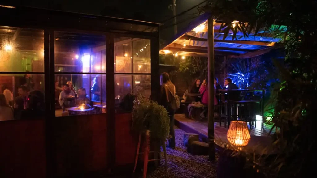 Nighttime outdoor bar/restaurant with warm lighting; people seated inside a glass-walled area and on the deck outside.