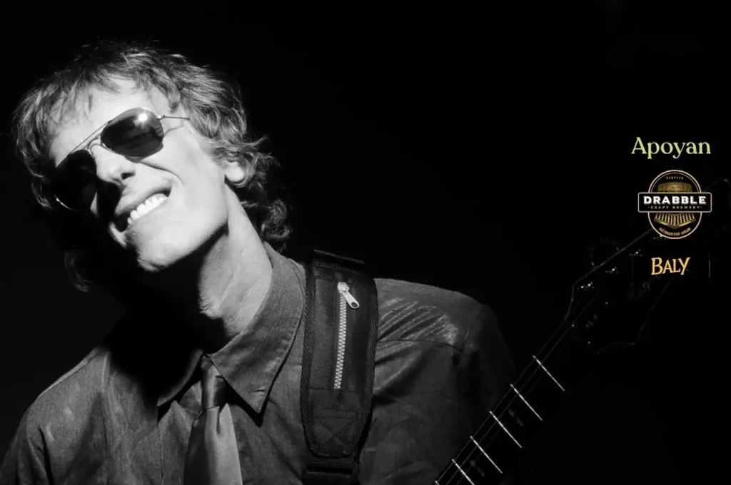 Black-and-white photo of a smiling guitarist wearing sunglasses, with sponsor logos on the right side on a black background.