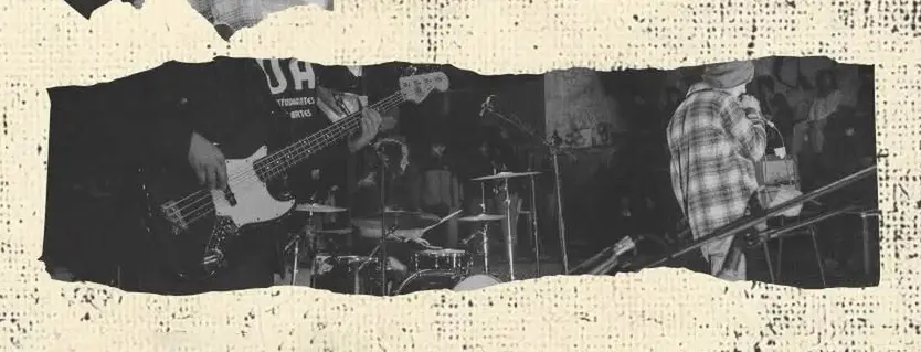 Black-and-white photo of a band playing on stage: bassist on the left, drummer center, singer on the right.