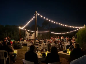 Outdoor night event with string lights, audience seated at tables watching a performer on a small stage.