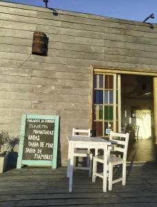 Rustic wooden café exterior with a chalkboard menu, outdoor white tables and chairs, and a rusty metal lantern mounted on the wall under a clear blue sky.