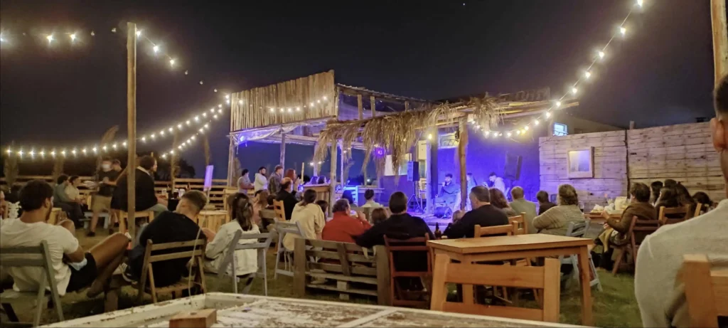 Outdoor nighttime concert at a rustic venue with string lights, a small stage, and audience seated at wooden tables under warm glow.