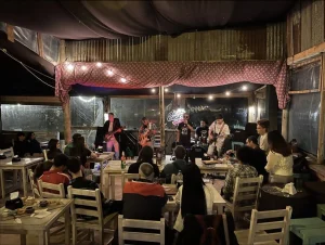 Casual restaurant with a small band performing on stage under pink fabric and warm string lights. Audience seated at wooden tables watching the musicians.