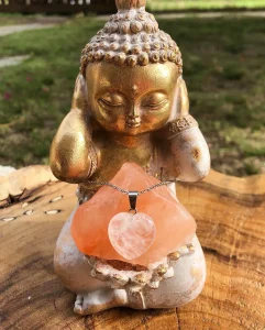 Gold Buddha statue figure wearing a pink heart-shaped crystal pendant, photographed outdoors on a wooden surface.