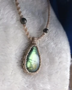 Beige knotted string necklace with a green iridescent teardrop pendant on a soft white surface.