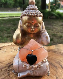 Small golden Buddha statue with hands cupping its head, placed on a wooden surface, adorned with a pink crystal and a black heart pendant necklace.