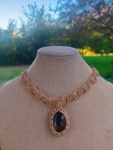 Macrame choker necklace with a large oval dark gemstone pendant displayed on a mannequin bust outdoors.